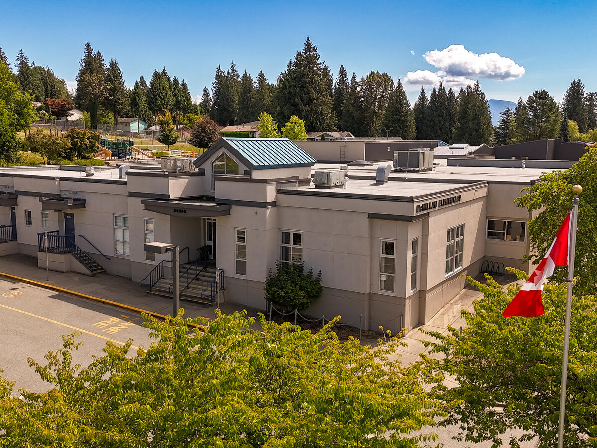 Exterior image of the entrance to an elementary school. Canadian flag on a pole out front of the school. Blue skies and green tree line in the background.