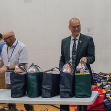 Three males standing at a table surrounded by food hampers, waiting to pass them to families