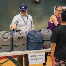 Two staff standing behind a table filled with backpacks as a student stands and waits to be handed one.