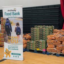Stacks of fresh produce in bags and boxes on crates with a marketing banner on display for "Archway"
